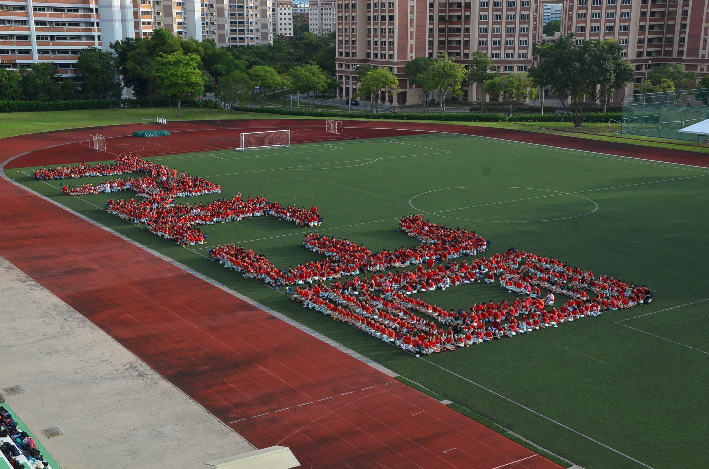 Hundreds of people in red shirts arranged on a green field to form large letter shapes, with a running track nearby.